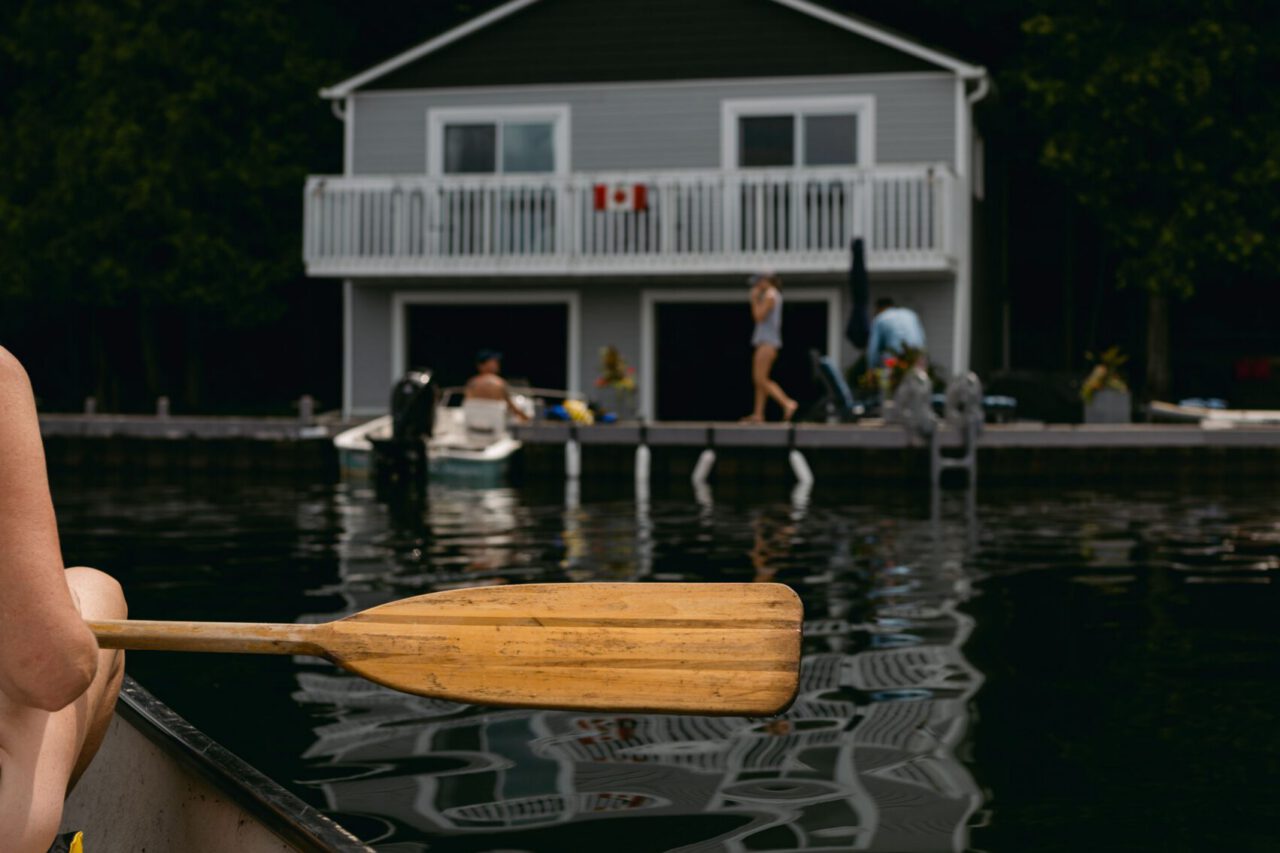 View past a canoe paddle toward a lakeside boathouse with people on the dock